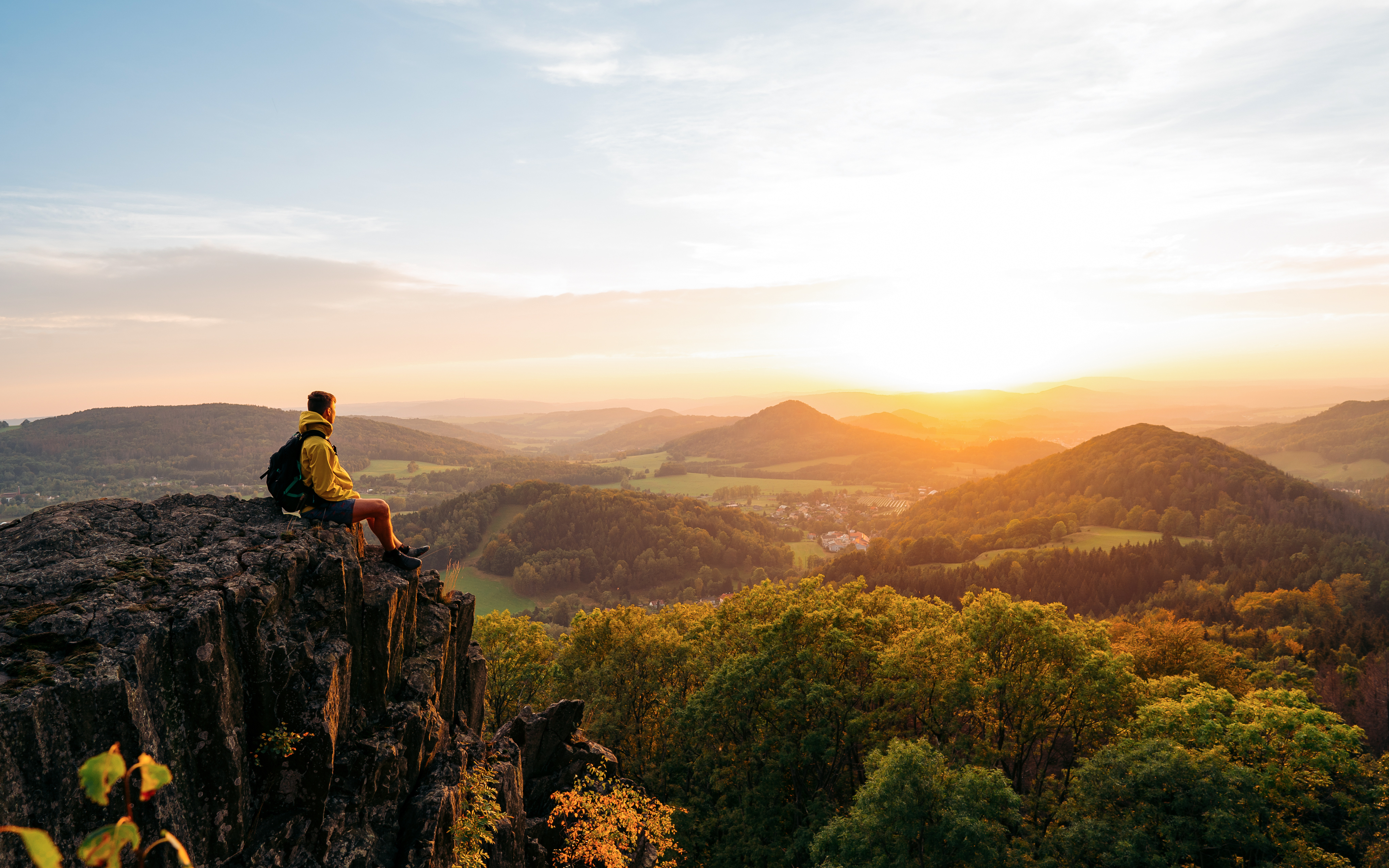 A person wearing a backpack sits on the edge of a rocky cliff, looking out over rolling green hills and forests at sunrise, with warm sunlight illuminating the valley below.