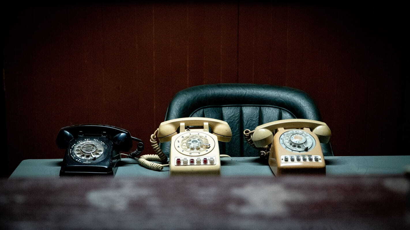 Three vintage rotary phones—black, cream, and beige—lined up on a desk in front of a green leather office chair against a dark wood-paneled wall.