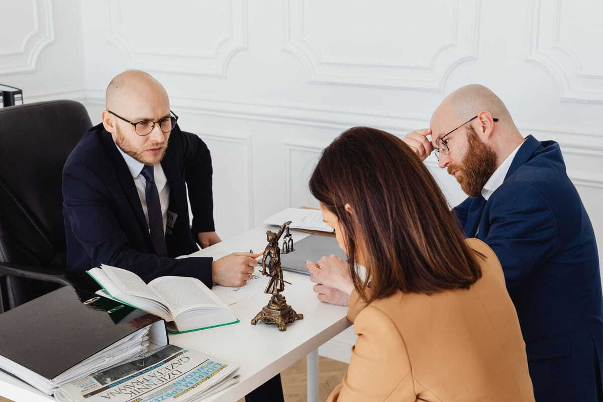 Two men in suits and a woman in a tan blazer sit around a desk during a legal consultation with documents, open books, and a small Statue of Justice on the table.   