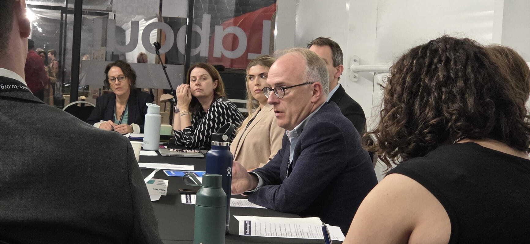 A panel discussion at a conference table, with a man in glasses speaking while other panelists listen attentively.   