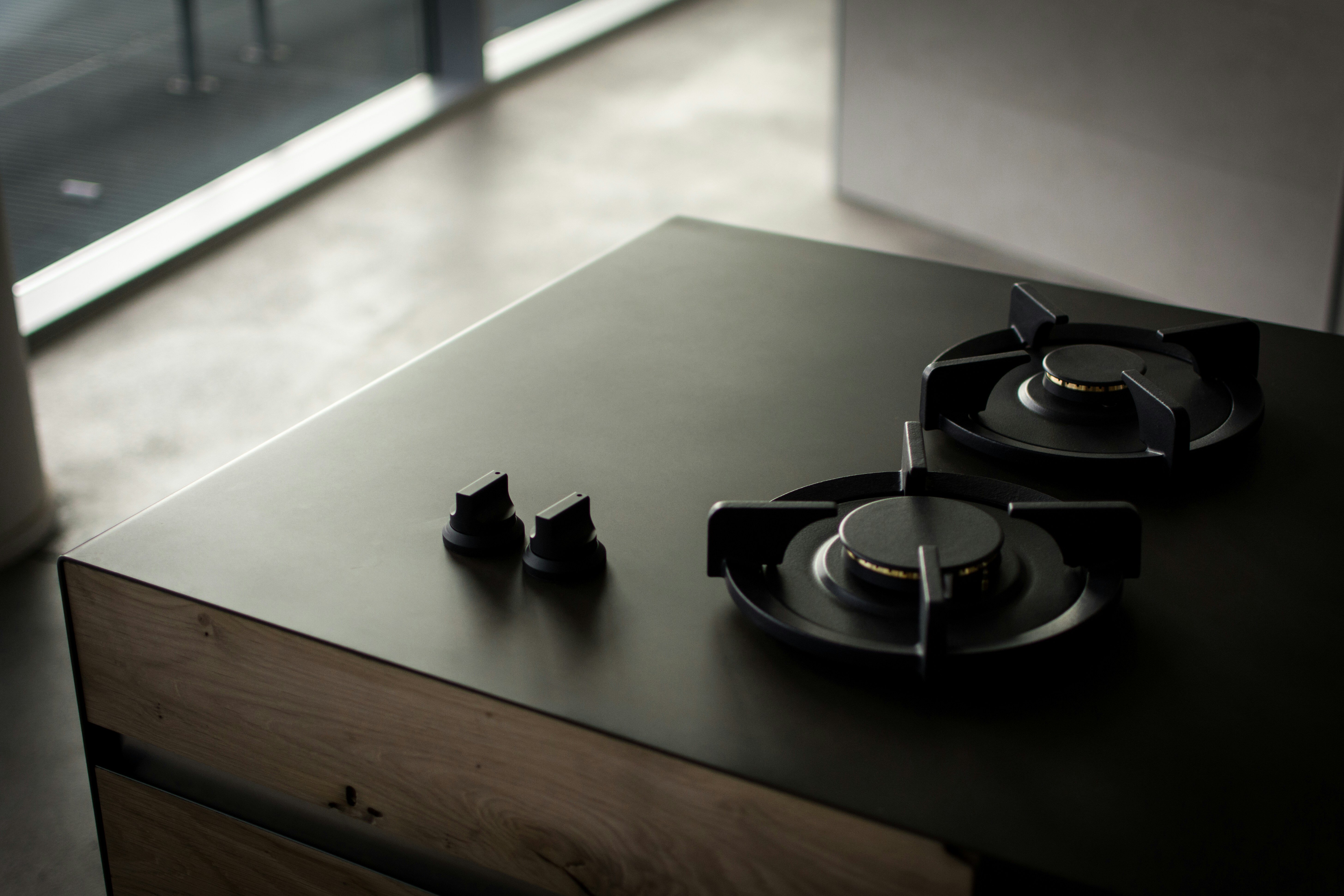 Downwards shot of a wooden unit with a smooth black countertop. On this, there are two gas hobs and two switches. In the background, there is a kitchen floor. 