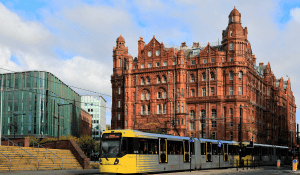 Yellow tram passing in front of a red-brick hotel, with modern glass buildings in the background.