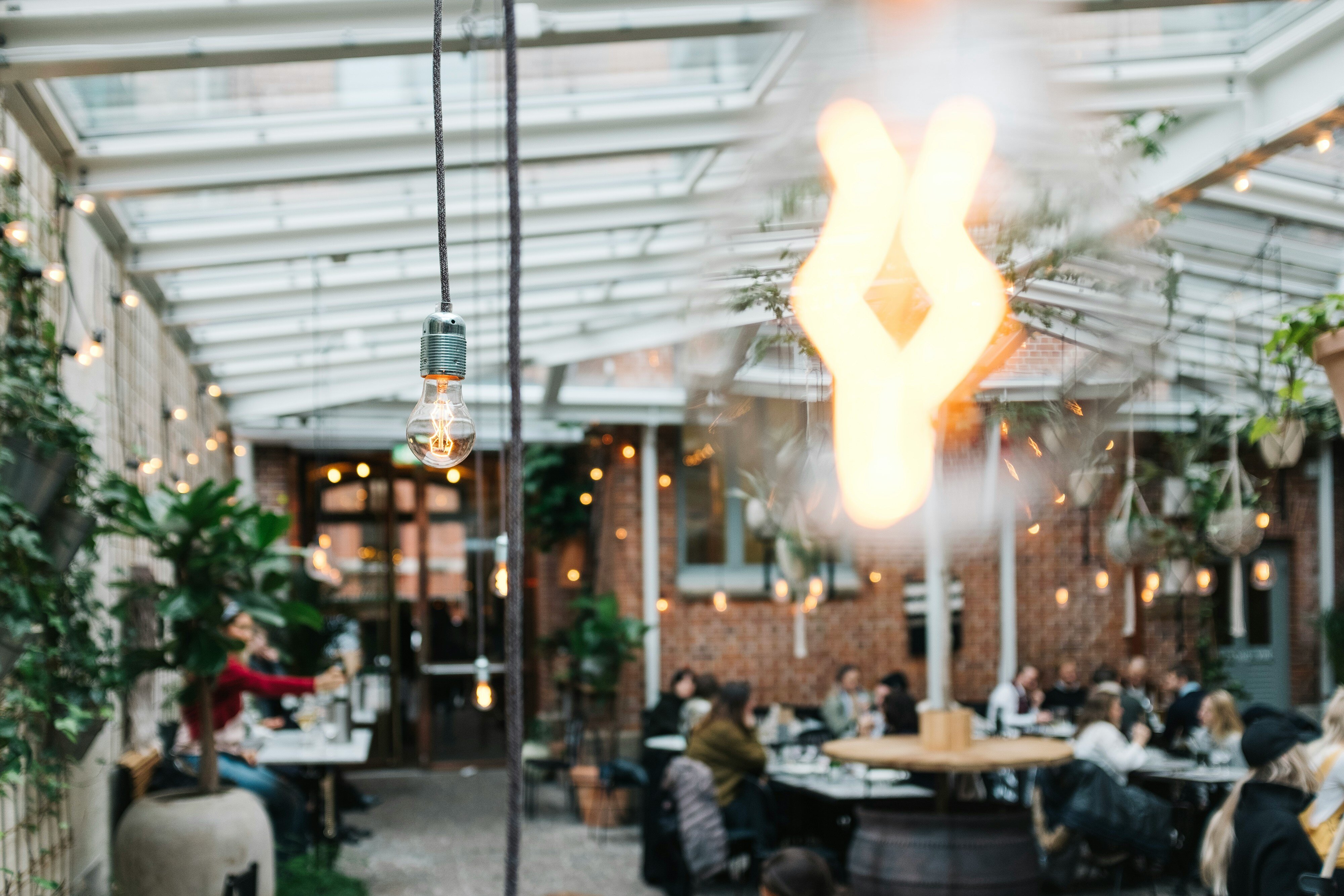 Image of a courtyard, sheltered by a glass-paned roof, with people sat down at tables chatting. The camera focuses on two free hanging lightbulbs in the foreground. 