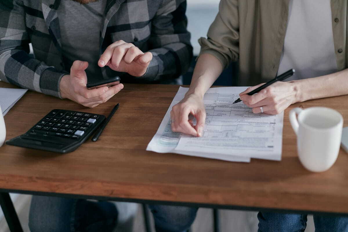 A close-up shot of a couple working together on their finances at a wooden table. On the left side of the frame, a man in a black and grey plaid button-up shirt and blue jeans holds a smartphone with both hands, looking down at the screen, with his left hand’s index finger touching the screen and his right hand's fingers holding the phone. Next to him is a black solar calculator with a black pen resting nearby. On the right side, a woman in an olive green button-up shirt with rolled-up sleeves and blue jeans, with a gold wedding band and a large diamond ring on her left hand, is holding a black pen in her right hand and pointing to a printed paper document with her left index finger as she marks on it. The couple is at a wooden table with printed financial forms, two white coffee mugs, a pencil, and a black solar calculator.