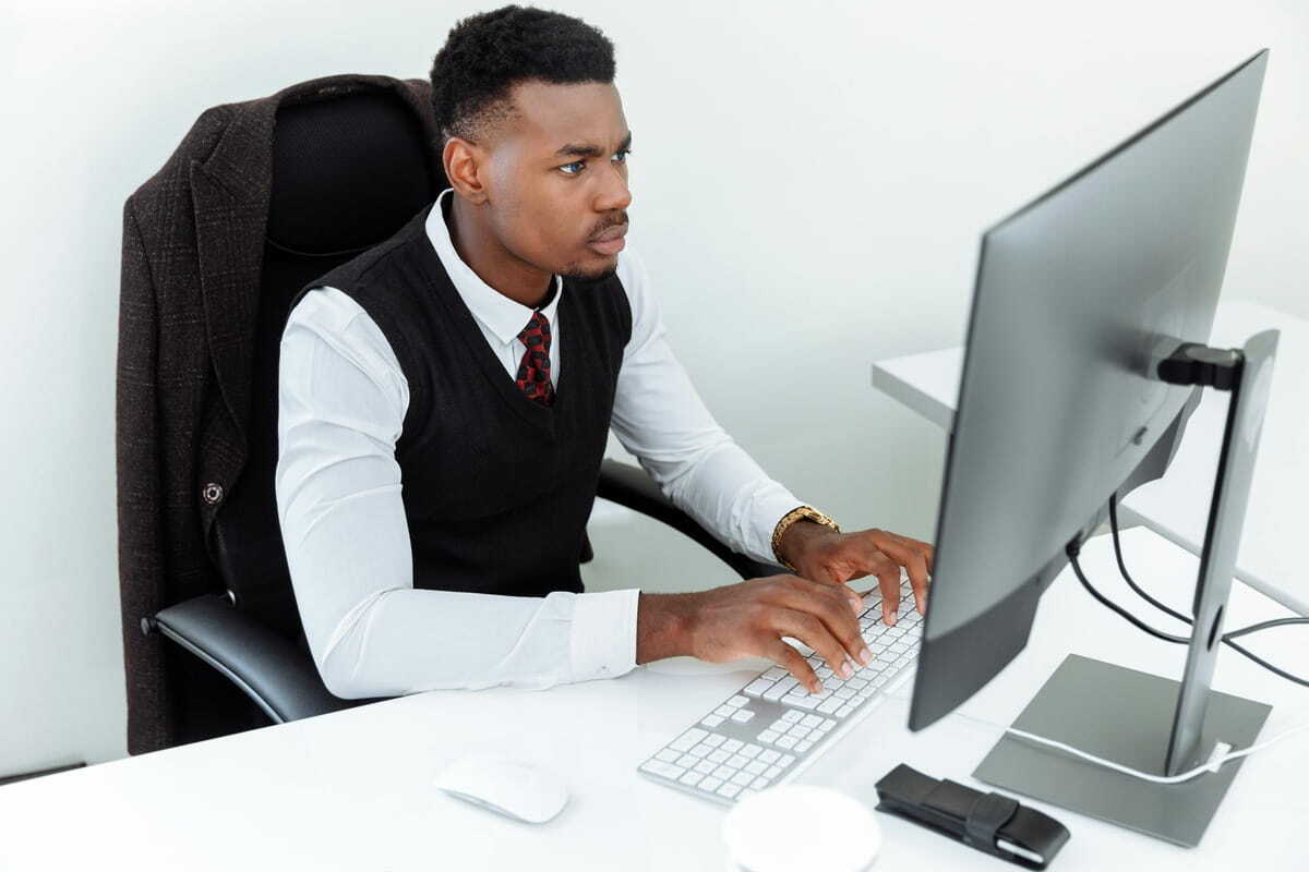 A man in a vest and tie works intently at a computer in a bright, modern office.