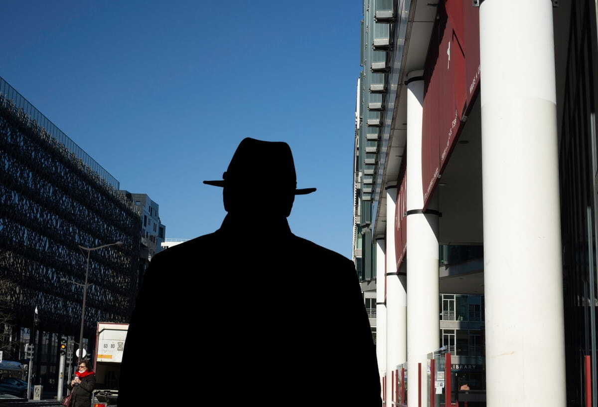 Silhouette of a person wearing a fedora hat and coat, walking down a sunny city street flanked by modern buildings and large white pillars.