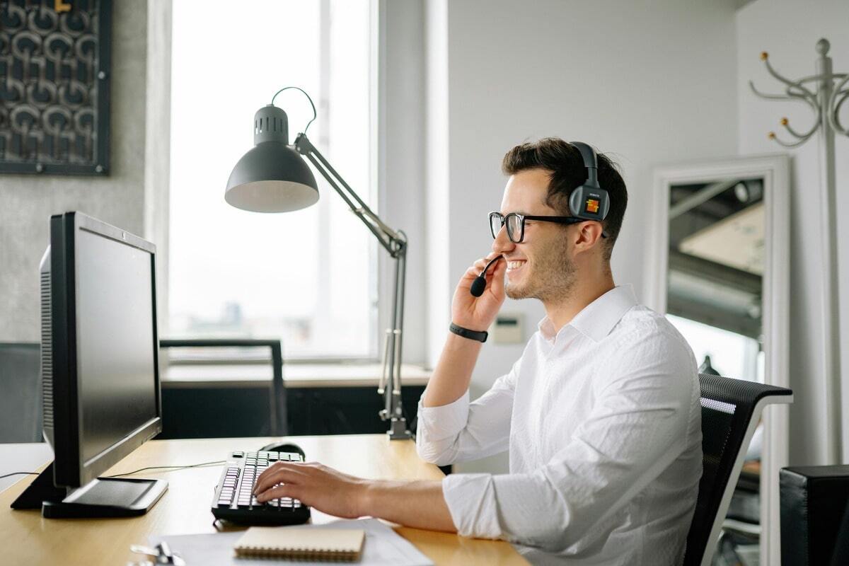 A smiling man wearing a white collared shirt, black-rimmed glasses, and a headset with a microphone sits at a desk, typing on a keyboard and looking at a computer monitor. He appears to be working in customer support or a call center. A black desk lamp is visible above the desk, and a bright window is in the background.