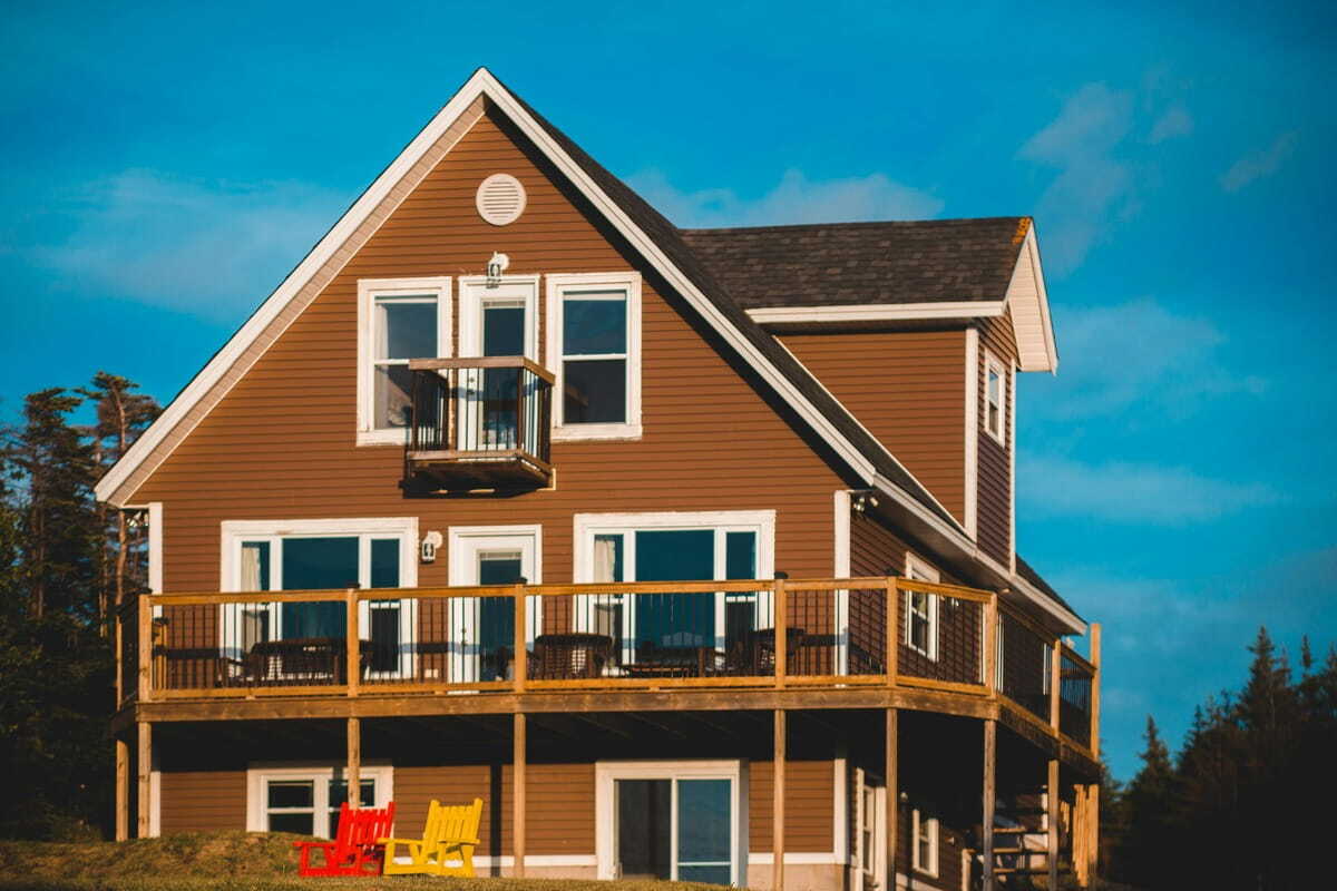 A brown two-story cottage with a large wooden wrap-around deck and a small upper balcony, set on a grassy hill under a blue sky.