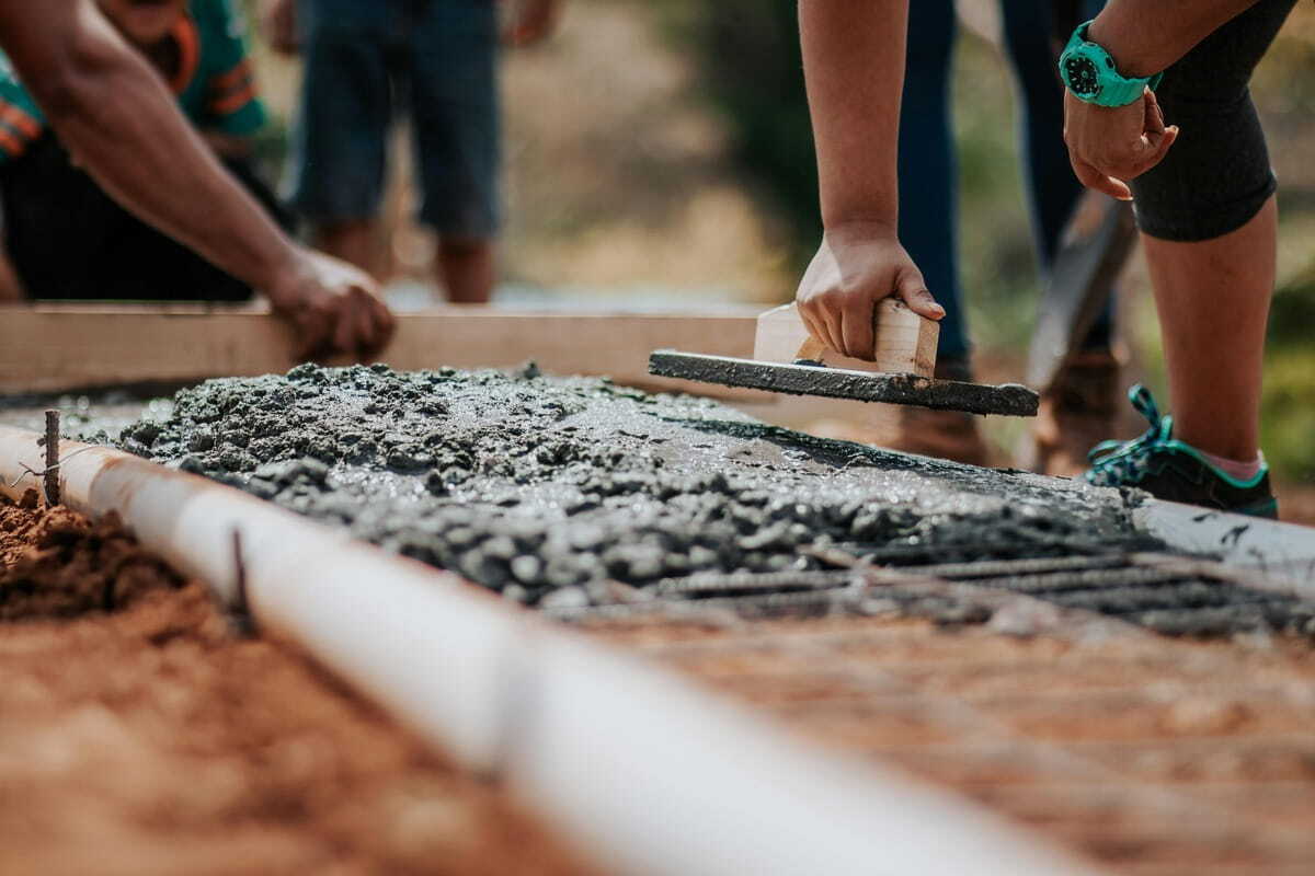 A medium close-up shows a person, from the waist down, and wearing a teal watch on their left wrist, actively smoothing a slab of fresh, grey, gravelly concrete using a trowel. To the right of the person, is a close view of fresh, grey concrete, full of gravel with two metal pipes running through it, and below it, is reddish, loose dirt. The background of the photo is blurry, but a person is standing to the right of the person smoothing the concrete. To the left is another person, from the waist up, bending over. Farther in the background is a blurry figure wearing blue shorts. The lighting in the photo is sunny, and bright.