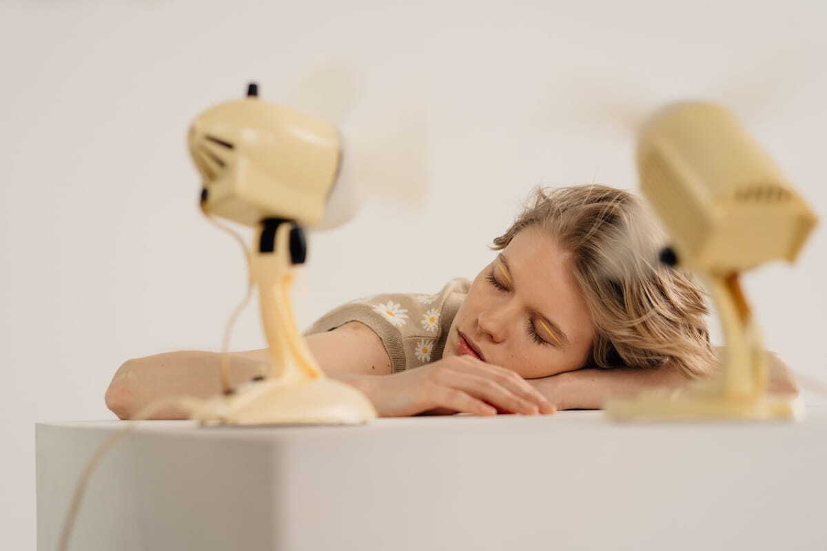 A person resting their head on a desk with eyes closed, positioned between two retro yellow electric fans blowing air toward them.