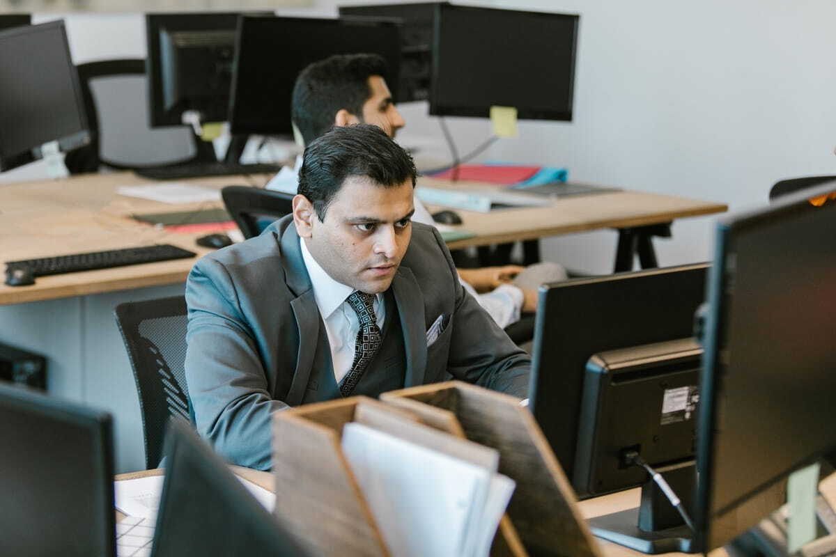 A man in a grey suit looks intently at a computer screen while working at his desk in an open-plan office.