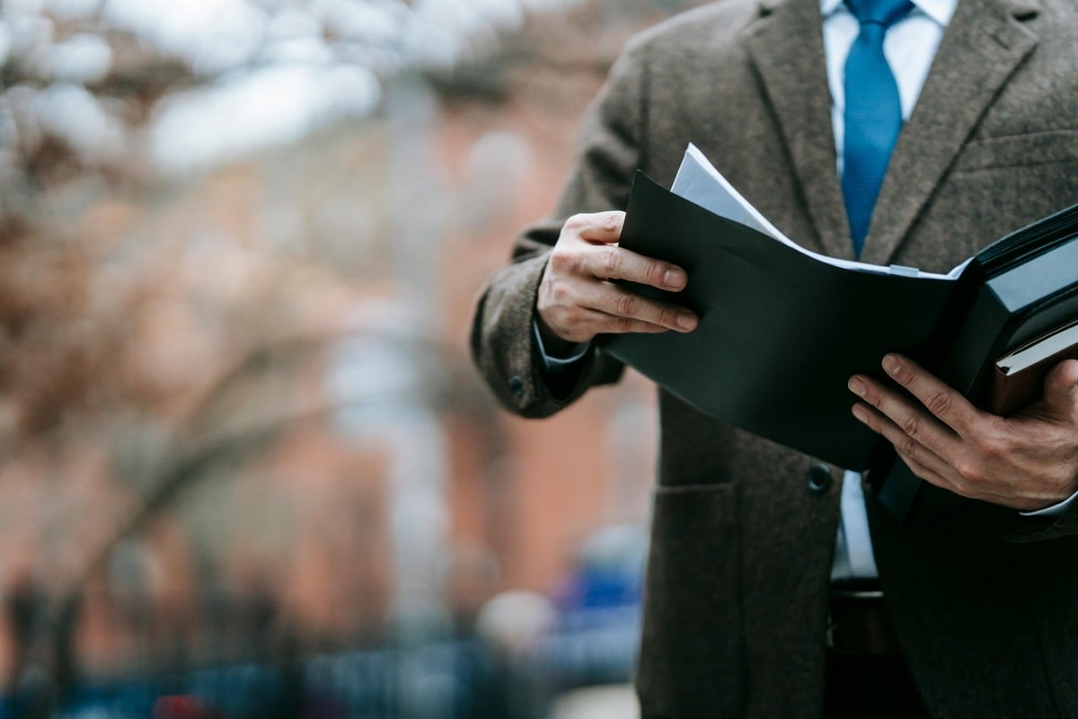 Close-up of a person in a brown tweed blazer, white shirt, and blue tie, holding open a black folder with documents outdoors.