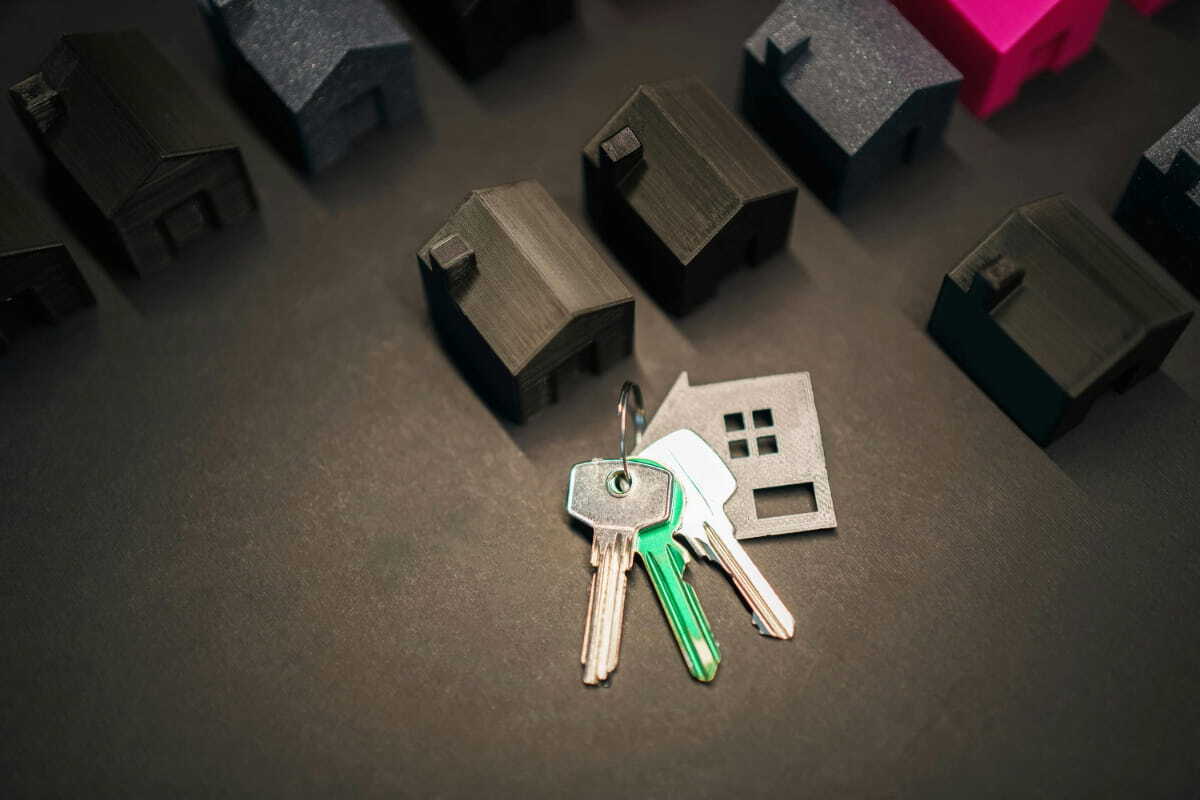 An overhead, close-up shot of a set of keys on a keyring, including a green key and a grey, house-shaped keychain. The keys are lying on a dark grey surface surrounded by small, 3D-printed model houses. Most of the houses are black or dark grey, with one vibrant pink house visible in the upper right background.