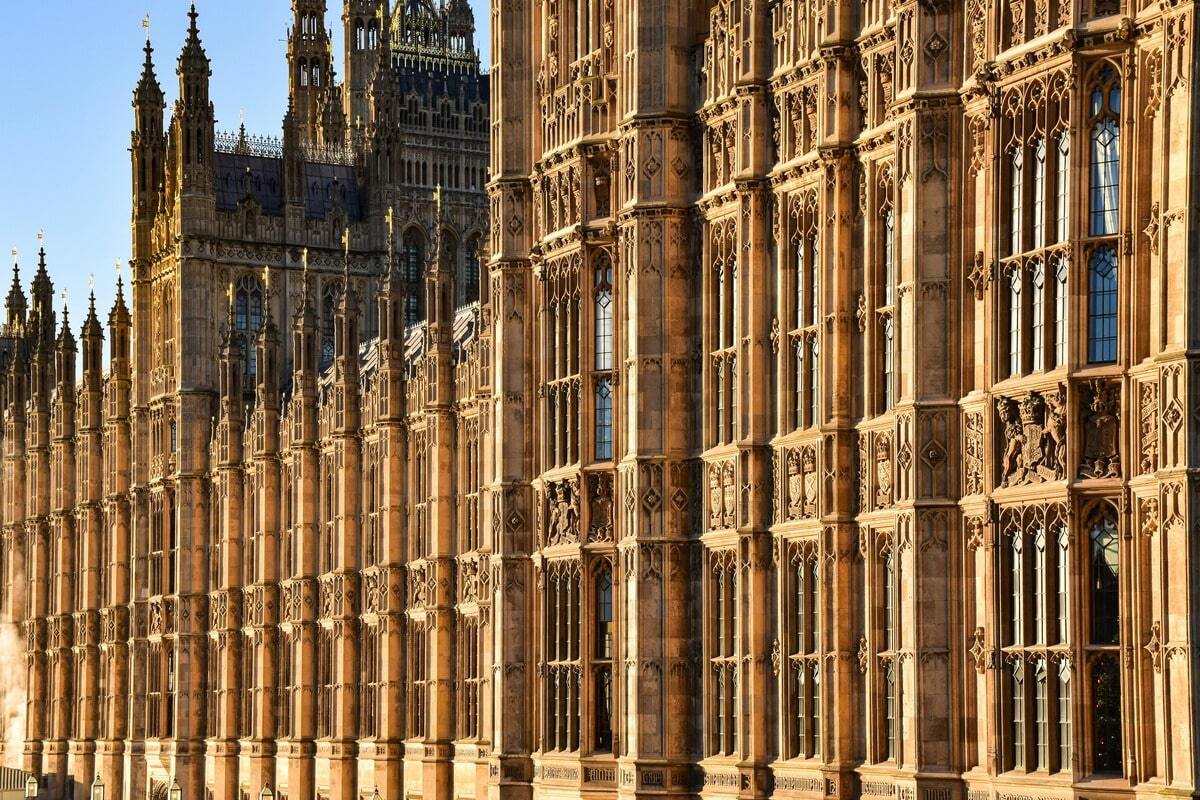 An architectural detail shot of the Palace of Westminster, showcasing its intricate Gothic Revival facade under a bright blue sky. The warm, honey-colored stone and ornate details of the building's towers and windows are highlighted by the sunlight.