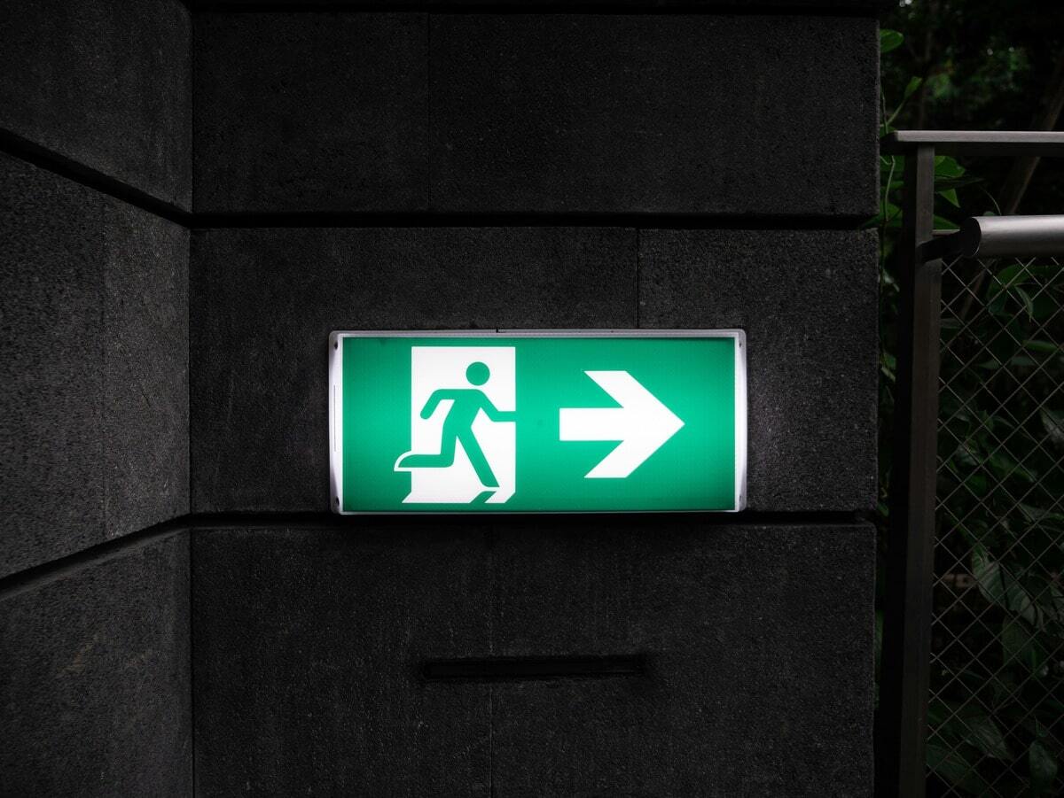 Illuminated green emergency exit sign on a dark wall, showing a person running through a door and an arrow pointing to the right, representing the exit of agency negotiators.