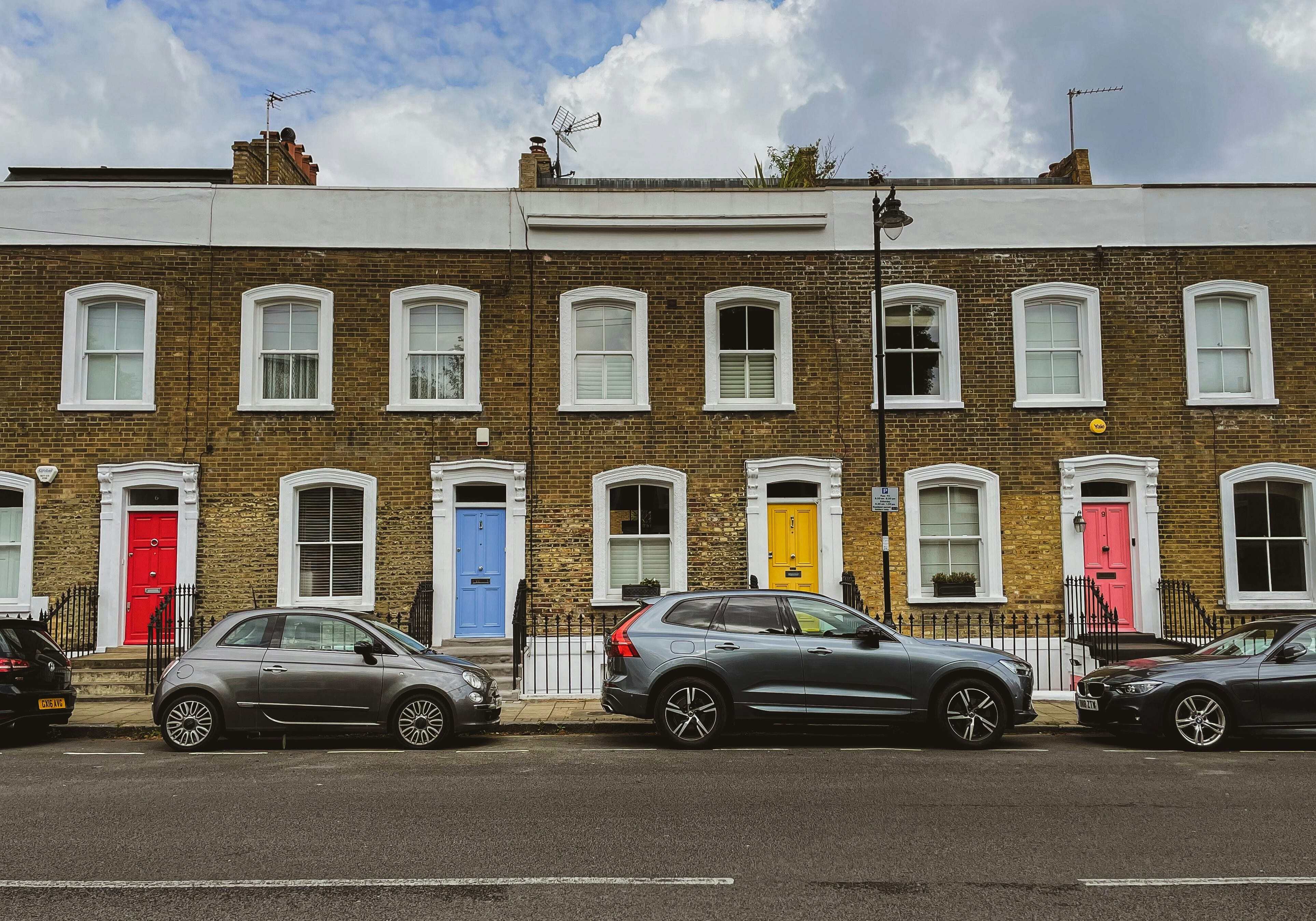 Row of traditional brick terraced houses with white-trimmed windows and brightly colored front doors (red, blue, yellow, pink) along a residential street, with parked cars in front under a partly cloudy sky.
