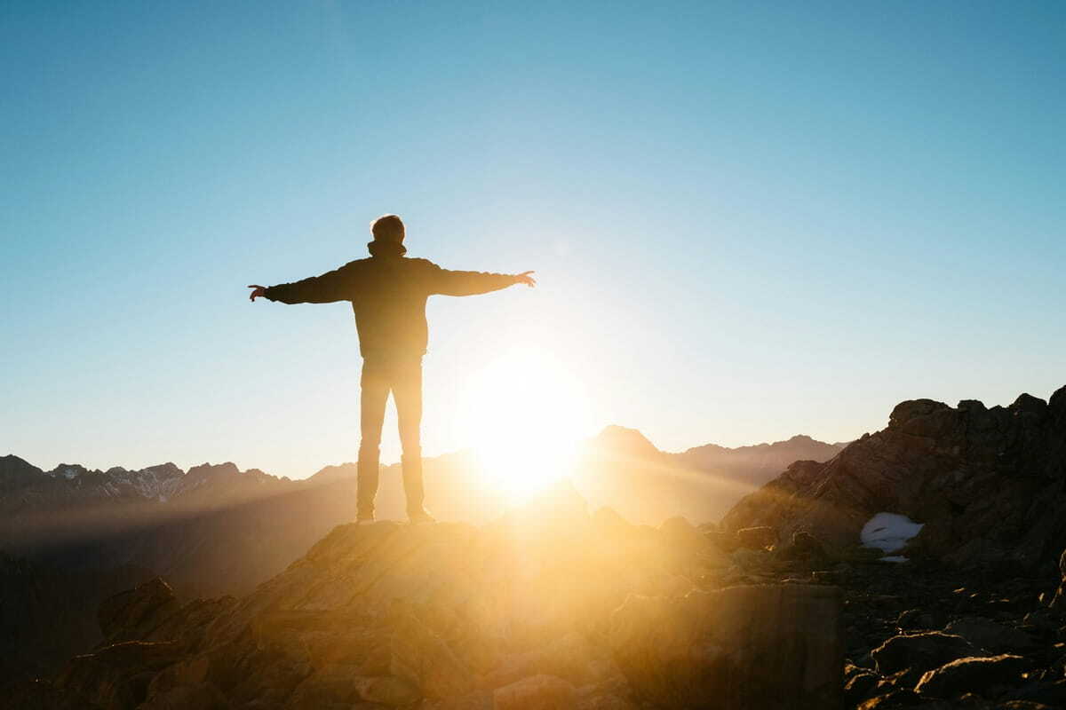 Silhouette of a person standing on a rocky mountain summit with arms open as the sun rises behind them.