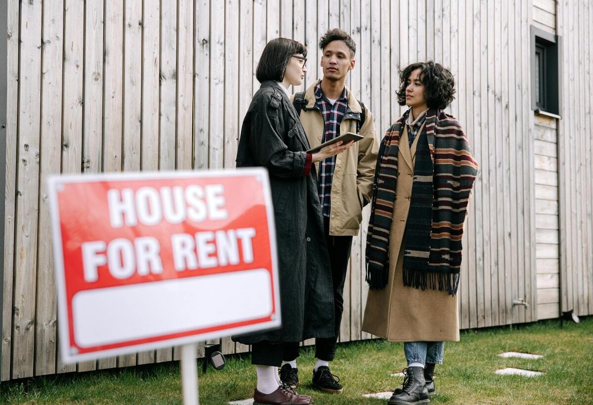  A group of three young adults stands outside a house with a "HOUSE FOR RENT" sign in the foreground. One person in a dark coat holds a tablet and speaks with the other two, who are dressed in a plaid shirt and a long striped scarf with a tan coat, respectively. The group appears to be discussing the rental property.