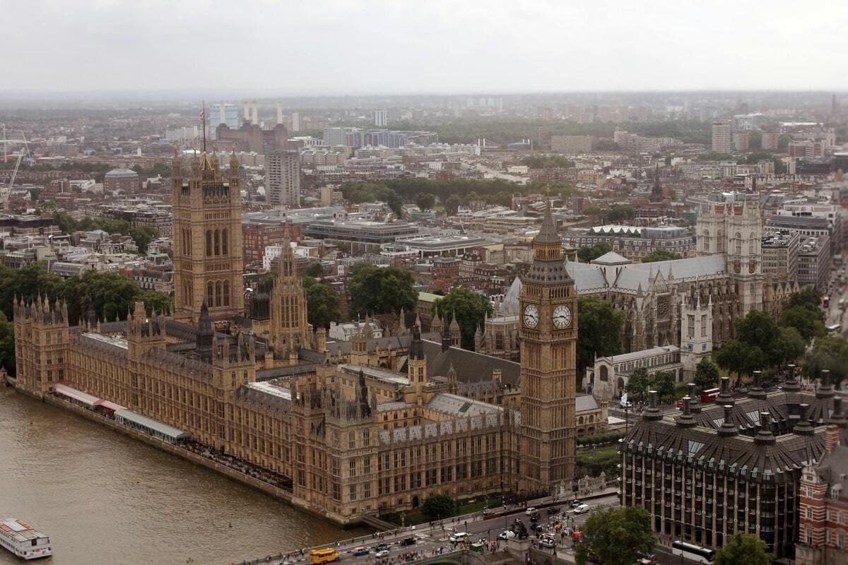 An aerial shot shows the Houses of Parliament and Big Ben in London, with the River Thames in the foreground. The Gothic Revival architecture of the Parliament building dominates the left and center of the frame, stretching along the riverbank. Big Ben, with its clock face visible, stands tall to the right of the Parliament building. Westminster Abbey is also visible to the right of Big Ben. The city extends into the hazy background, with a mix of historical and modern buildings under an overcast sky. A white boat is visible on the River Thames in the bottom left, and traffic can be seen on the road running parallel to the river.