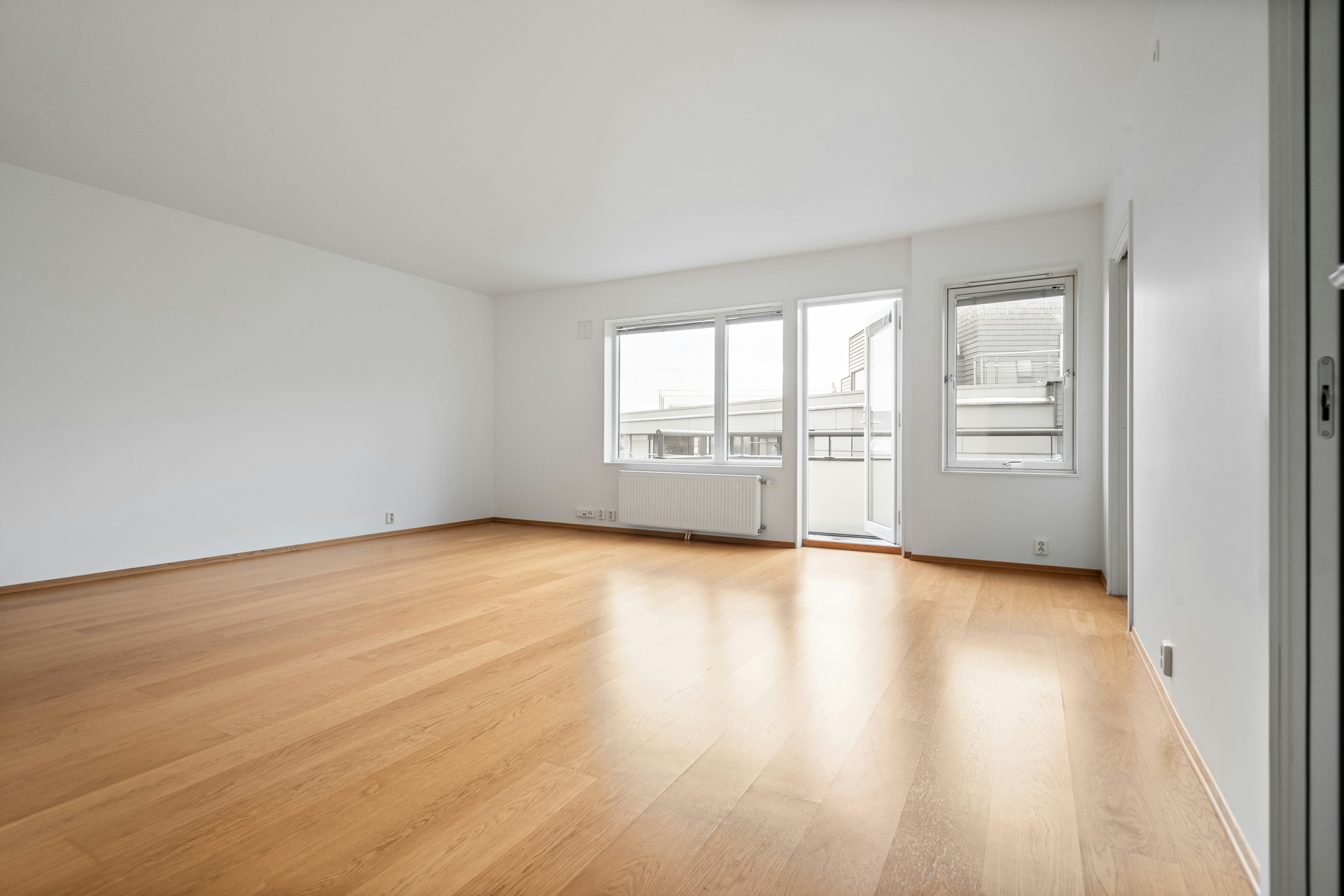 An image of an empty flat with a yellow birch wooden floor. The door at the far end of the flat is open but the windows are closed with blinds un-drawn. The walls, plug sockets and fittings are all white. 